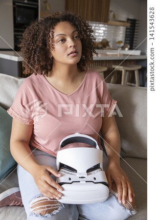 African American woman holding VR headset on lap, gazing upward in living room near kitchen island African American woman holding VR headset on lap, gazing upward in living room near kitchen island 141543628