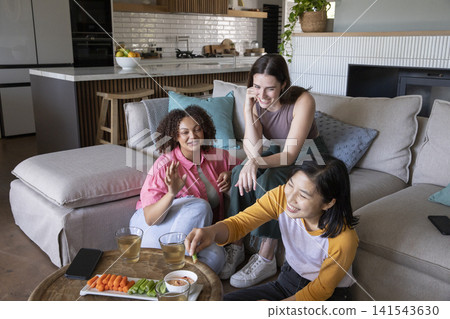 Diverse female friends sitting on sofa in living room dipping veggies in dip near coffee table Diverse female friends sitting on sofa in living room dipping veggies in dip near coffee table 141543630