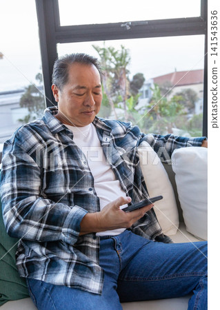 Asian man sitting on sofa at home near large window holding smartphone in plaid shirt Asian man sitting on sofa at home near large window holding smartphone in plaid shirt 141543636