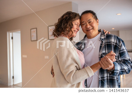 Diverse senior couple dancing in open plan living area with framed prints and kitchen island Diverse senior couple dancing in open plan living area with framed prints and kitchen island 141543637
