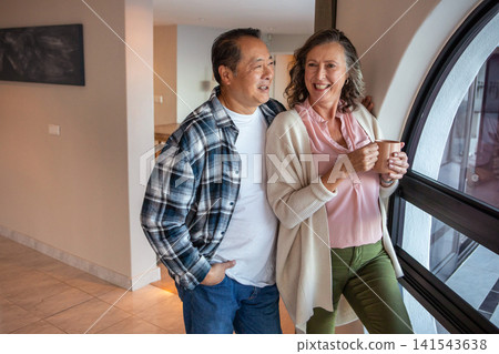 Diverse senior couple standing by window at home holding ceramic mug and gazing outside Diverse senior couple standing by window at home holding ceramic mug and gazing outside 141543638
