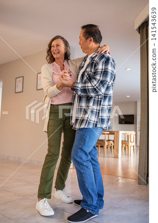 Diverse couple dancing joyfully in open plan living area near wooden table and framed prints Diverse couple dancing joyfully in open plan living area near wooden table and framed prints 141543639