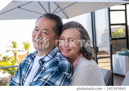 Diverse couple hugging on terrace loveseat under patio umbrella overlooking garden 141543641
