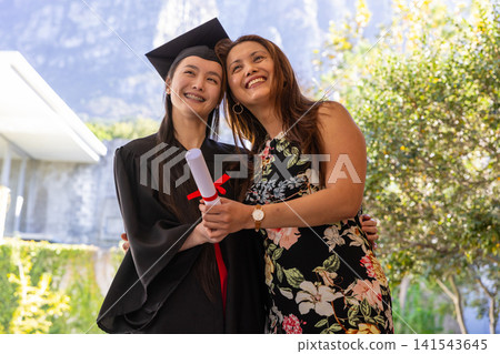 Mother and daughter posing on campus wearing graduation cap and gown and holding diploma degree Mother and daughter posing on campus wearing graduation cap and gown and holding diploma degree 141543645
