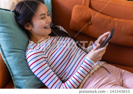 Asian woman reclining on couch using smartphone and resting head on teal pillow Asian woman reclining on couch using smartphone and resting head on teal pillow 141543653