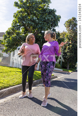 Diverse female friends walking along sidewalk holding bottle, wearing fitness trackers Diverse female friends walking along sidewalk holding bottle, wearing fitness trackers 141543654