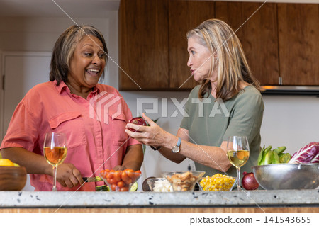 Diverse senior friends standing at marble kitchen island, slicing cucumbers, holding wine Diverse senior friends standing at marble kitchen island, slicing cucumbers, holding wine 141543655