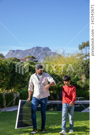 Indian father son walking on green lawn beside pool carrying solar panel with mountain backdrop Indian father son walking on green lawn beside pool carrying solar panel with mountain backdrop 141543671