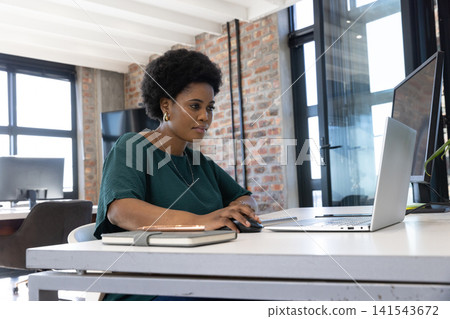 Mid adult african american woman working at desk in office using laptop and wireless mouse Mid adult african american woman working at desk in office using laptop and wireless mouse 141543672