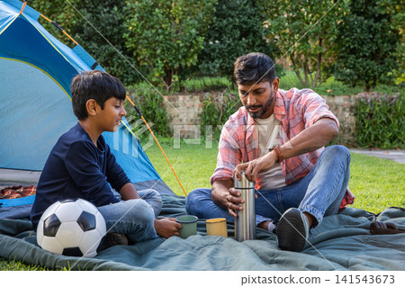 Indian father with son sitting cross-legged on lawn in front of blue tent, pouring from thermos Indian father with son sitting cross-legged on lawn in front of blue tent, pouring from thermos 141543673