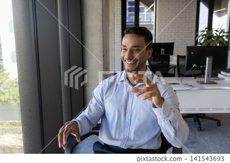 Male office worker gesturing and sitting in office chair beside window at desk with dual monitors Male office worker gesturing and sitting in office chair beside window at desk with dual monitors 141543693