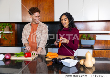 Diverse couple chopping vegetables on wooden cutting board and peeling carrot at kitchen Diverse couple chopping vegetables on wooden cutting board and peeling carrot at kitchen 141543709