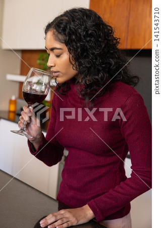 Indian woman smelling red wine in glass at kitchen counter by potted plant and bottle Indian woman smelling red wine in glass at kitchen counter by potted plant and bottle 141543710