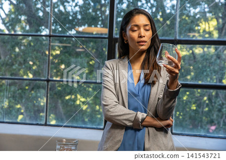 Asian woman standing by window at home, holding stemless glass beside glass jar 141543721