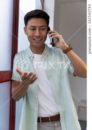 Asian man leaning on panel in hallway, talking on smartphone and gesturing Asian man leaning on panel in hallway, talking on smartphone and gesturing 141543743