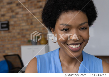 African American woman wearing blue blouse smiling while sitting at small office with whiteboard African American woman wearing blue blouse smiling while sitting at small office with whiteboard 141543744