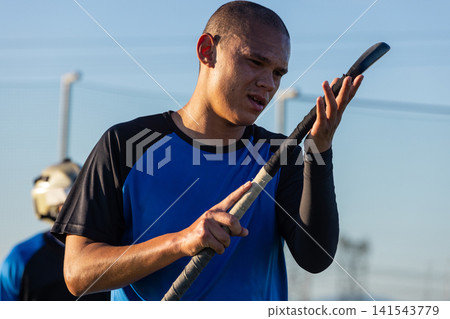 Hispanic male athlete inspecting taped hockey stick at fence next to goal net 141543779