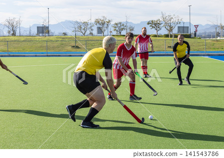 Male players dribbling white ball with hockey sticks on green turf while competing near fence 141543786