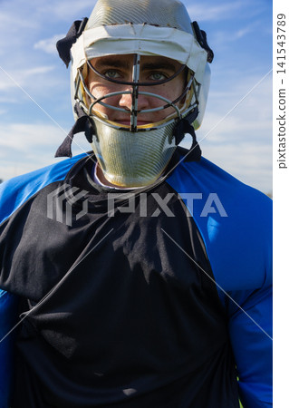 Teenage boy standing on lacrosse field in goalie helmet and padded jersey under blue sky 141543789