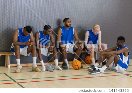 Male teammates resting on wooden bench, polished hardwood court floor at gym around basketballs Male teammates resting on wooden bench, polished hardwood court floor at gym around basketballs 141543823