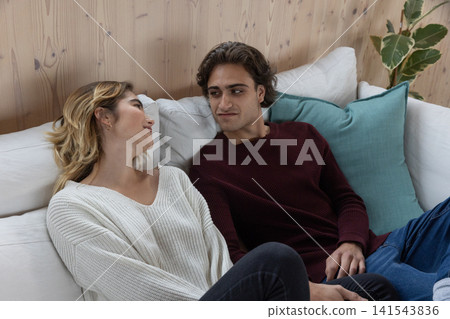 Couple reclining on white sofa at home with wood-paneled wall, exchanging smiles and eye contact Couple reclining on white sofa at home with wood-paneled wall, exchanging smiles and eye contact 141543836
