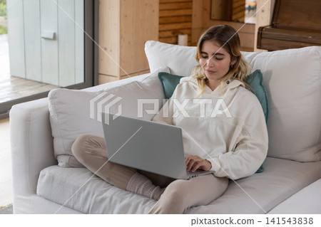 Woman typing on silver laptop while sitting cross-legged on cushioned sofa in wood-paneled cabin Woman typing on silver laptop while sitting cross-legged on cushioned sofa in wood-paneled cabin 141543838