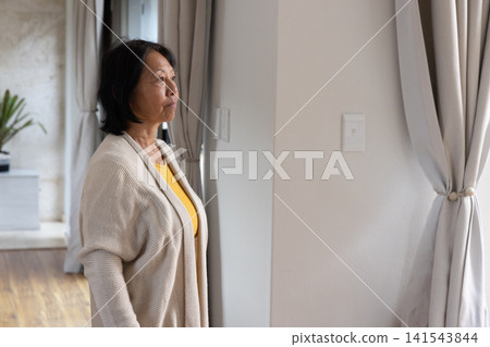 Senior Asian woman standing in living area gazing through curtains at window beside potted plant Senior Asian woman standing in living area gazing through curtains at window beside potted plant 141543844