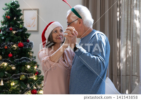 Senior couple dancing in living room with Christmas tree wearing Santa hat and antler headband Senior couple dancing in living room with Christmas tree wearing Santa hat and antler headband 141543858