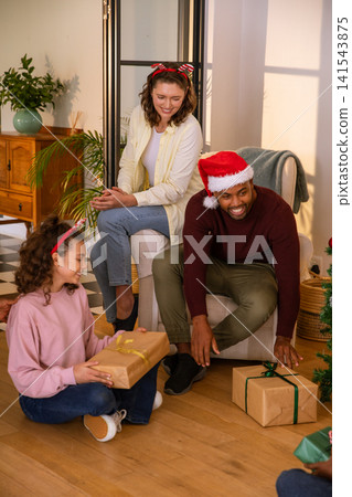 Diverse family wearing santa hats gathering around wrapped gifts in living room Diverse family wearing santa hats gathering around wrapped gifts in living room 141543875
