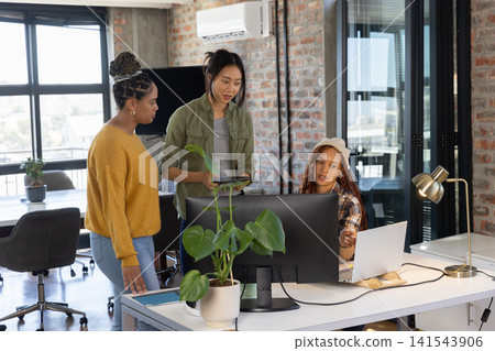 Diverse female coworkers collaborating around desk in modern office with monitor, laptop, tablet Diverse female coworkers collaborating around desk in modern office with monitor, laptop, tablet 141543906