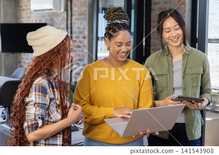 Diverse female coworkers collaborating on silver laptop and black tablet in brick-walled office Diverse female coworkers collaborating on silver laptop and black tablet in brick-walled office 141543908