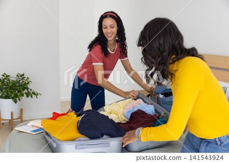Diverse mom and teenage daughter packing gray suitcase with folded clothes on bed near plant Diverse mom and teenage daughter packing gray suitcase with folded clothes on bed near plant 141543924