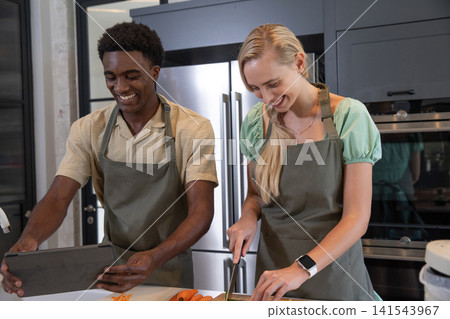 Diverse couple prepping meal at kitchen island using tablet, slicing carrots on cutting board Diverse couple prepping meal at kitchen island using tablet, slicing carrots on cutting board 141543967