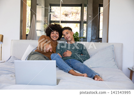 Diverse male partners using silver laptop while sitting on king-size bed in modern bedroom Diverse male partners using silver laptop while sitting on king-size bed in modern bedroom 141543995