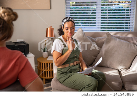 Diverse female counselor and client discussing therapy notes on sofa with paper, pen Diverse female counselor and client discussing therapy notes on sofa with paper, pen 141543999