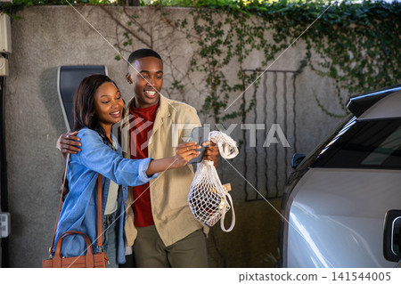 African American couple taking selfie with smartphone at carport holding grocery bag by charger 141544005