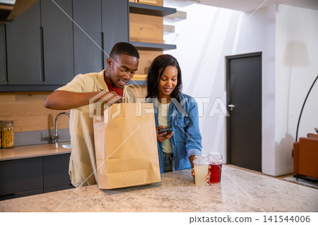 African American couple unpacking grocery bag on kitchen island checking phone and placing drinks African American couple unpacking grocery bag on kitchen island checking phone and placing drinks 141544006