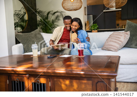 African American couple hugging and sharing takeout in home living room by coffee table with cups 141544017