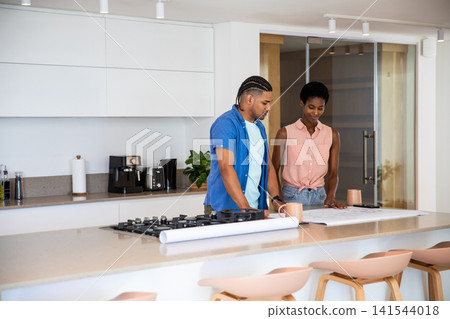 Diverse couple standing at kitchen island by gas cooktop examining blueprints holding coffee mugs Diverse couple standing at kitchen island by gas cooktop examining blueprints holding coffee mugs 141544018