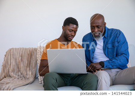 African american father and son sitting on sofa with beige throw pointing at laptop African american father and son sitting on sofa with beige throw pointing at laptop 141544023