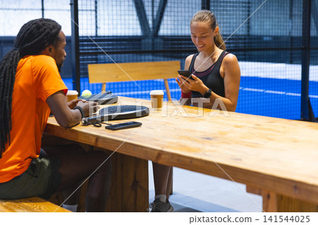 Diverse friends sitting at table by padel court with paddles, ball sipping coffee using smartphone Diverse friends sitting at table by padel court with paddles, ball sipping coffee using smartphone 141544025