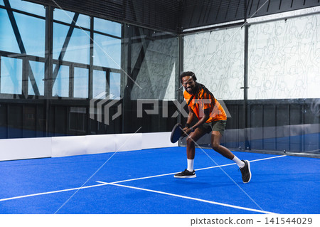 African American male athlete holding paddle and playing on blue court, glass walls and steel beams African American male athlete holding paddle and playing on blue court, glass walls and steel beams 141544029