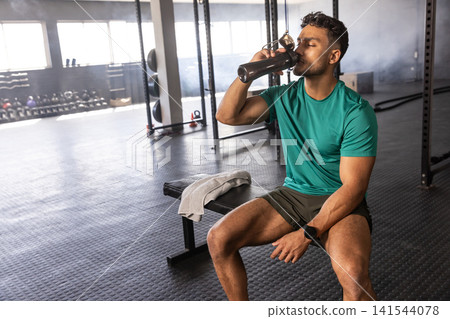 Asian male sitting on workout bench in gym drinking from shaker bottle beside gray towel Asian male sitting on workout bench in gym drinking from shaker bottle beside gray towel 141544078