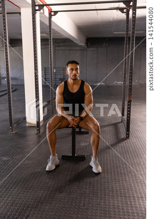 Hispanic male athlete sitting on bench beside squat rack with pull-up bars in well-equipped gym Hispanic male athlete sitting on bench beside squat rack with pull-up bars in well-equipped gym 141544080