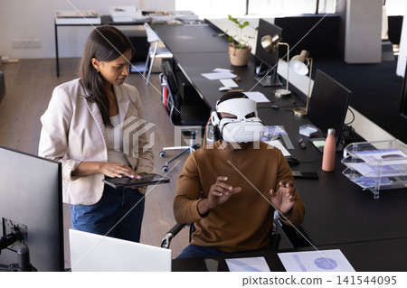 Diverse coworkers using vr headset and tablet at office desk with one in wheelchair Diverse coworkers using vr headset and tablet at office desk with one in wheelchair 141544095