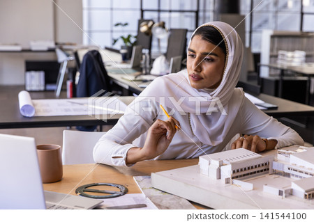 Asian indian woman wearing hijab examining model at studio desk with laptop and pencil, copy space Asian indian woman wearing hijab examining model at studio desk with laptop and pencil, copy space 141544100