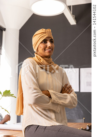 Indian woman leaning against desk in office studio holding blueprints next to pinned sketches Indian woman leaning against desk in office studio holding blueprints next to pinned sketches 141544102