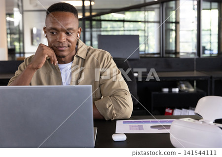 Mid adult african american man sitting at desk using laptop with chart, headset, copy space 141544111