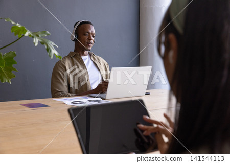 African american coworkers using laptop, tablet, headphones at table with plant, copy space 141544113