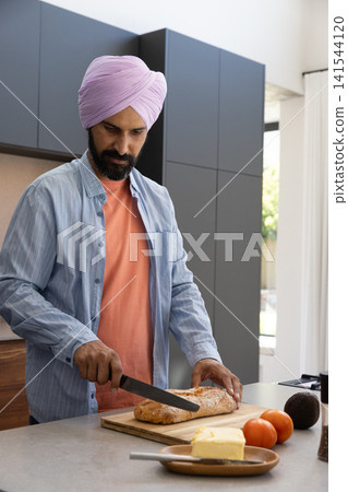 Indian man slicing crusty loaf on cutting board at kitchen island with tomatoes, avocado Indian man slicing crusty loaf on cutting board at kitchen island with tomatoes, avocado 141544120